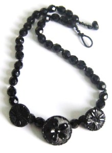 Image shows a necklace made from a strand of opaque faceted black glass beads of about 8mm. At the front are 3 large round buttons with a carved style flower motiff in the centre, made from black glass.