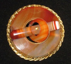 image shows a large antique brooch made from banded red agate. It is circular, with a hole in the centre. On top of this hole is another carved agate small circle and inside of this, a carved long oval straight piece. The whole brooch imitates a buckle. The brooch is set in gold, all around the rim.