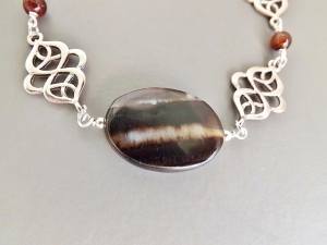 photo shows a close up of a necklace, with oval brown and cream banded agate cabochon bezel set, and two Celtic style knot patterns of about the same size (approx 4 cm) on either side.
