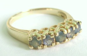 image shows a gold ring, set with five small round cut alexandrite gemstones, which are a weak misty pale teal-green colour