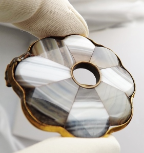 image shows a close up of some inlay work of a large roundish brooch; there a eight inlays of various types of banded agate all in brown black and white striped patterns, each of these inlays are flattened semi-teardrop shaped, and they are place in a way that is circular, almost creating a flower petal effect