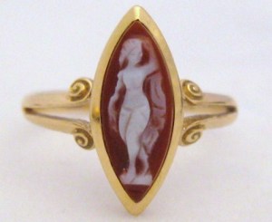 photo shows a close up of a gold ring. The central stone is a large marquise shaped cameo. This cameo has a red background and depicts a white figure of a naked woman holding some type of towel or clock behind her, and is quite crudely carved.