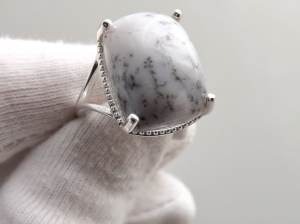 image shows a white gloved hand holding a silver ring to the camera, the ring is set with a large rectangular cabochon cut white dendritic agate gemstone, opaque white in colour, with dark green-grey fern like pattern inclusions (dendrils)