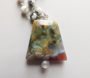 photo shows a close up of an irregular rectangle type shape agate pendant which is beautoful patterns with bands and swirls in various white, cream and pale orange and green colours