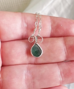 photo shows a semi teardrop shaped emerald, a flat cabochon design, set into hand textured sterling silver bezel style pendant with two loops at the top where the chain it comes with goes on.